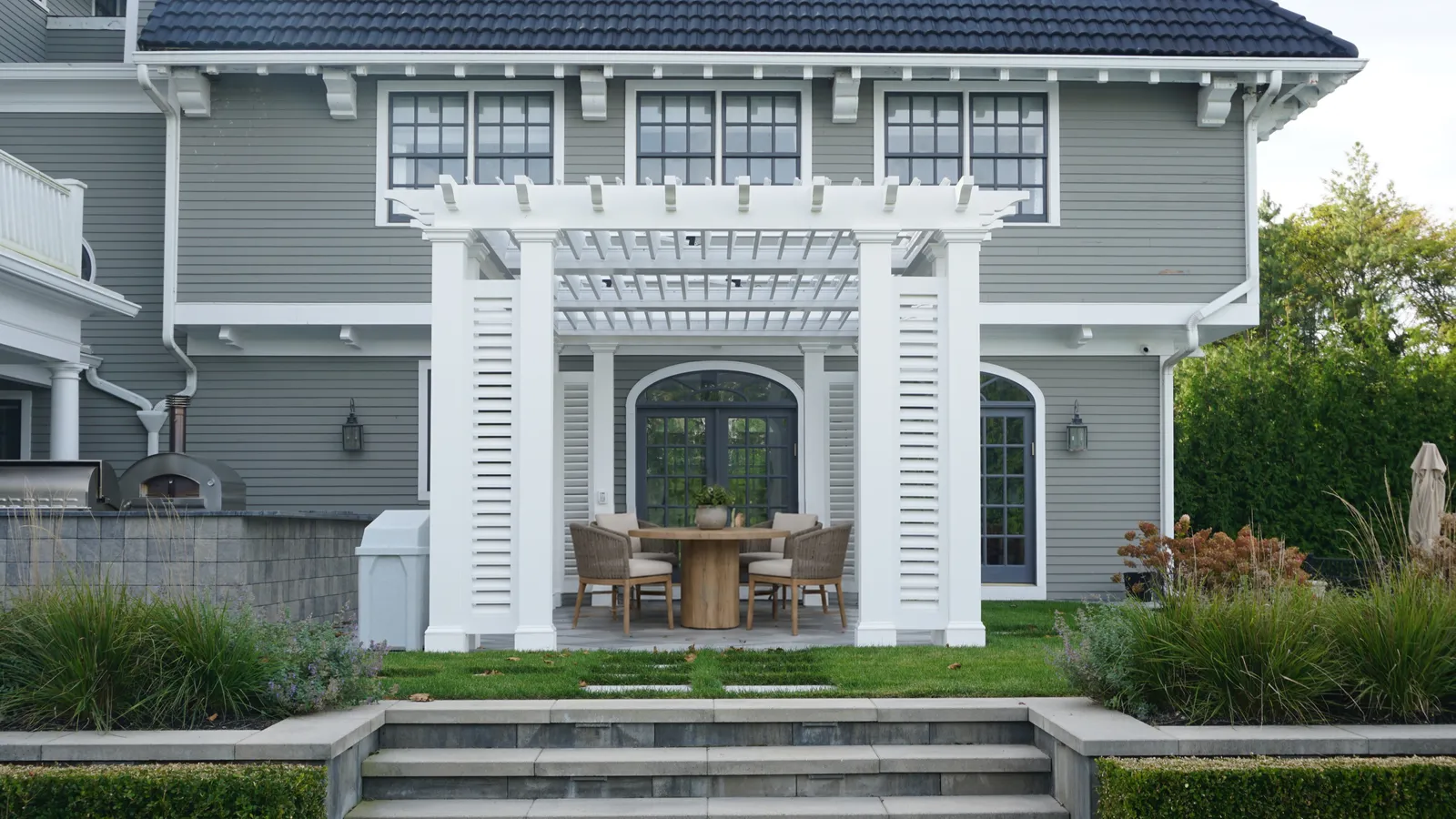 White pergola with louvered columns over a dining terrace, centered on stone steps up from the lawn